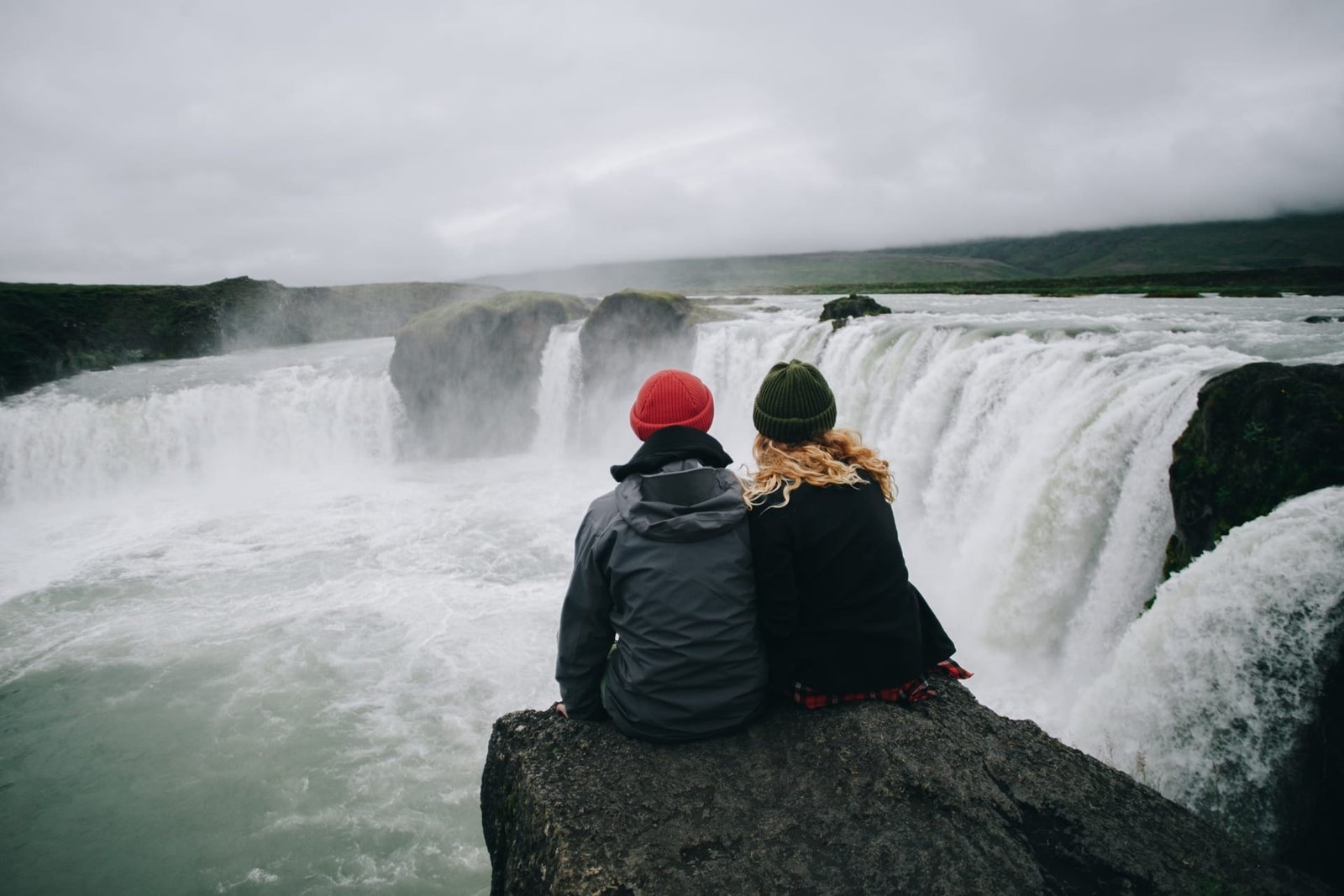 Two friends in knit hats sitting on a rock above a wide, roaring Icelandic waterfall