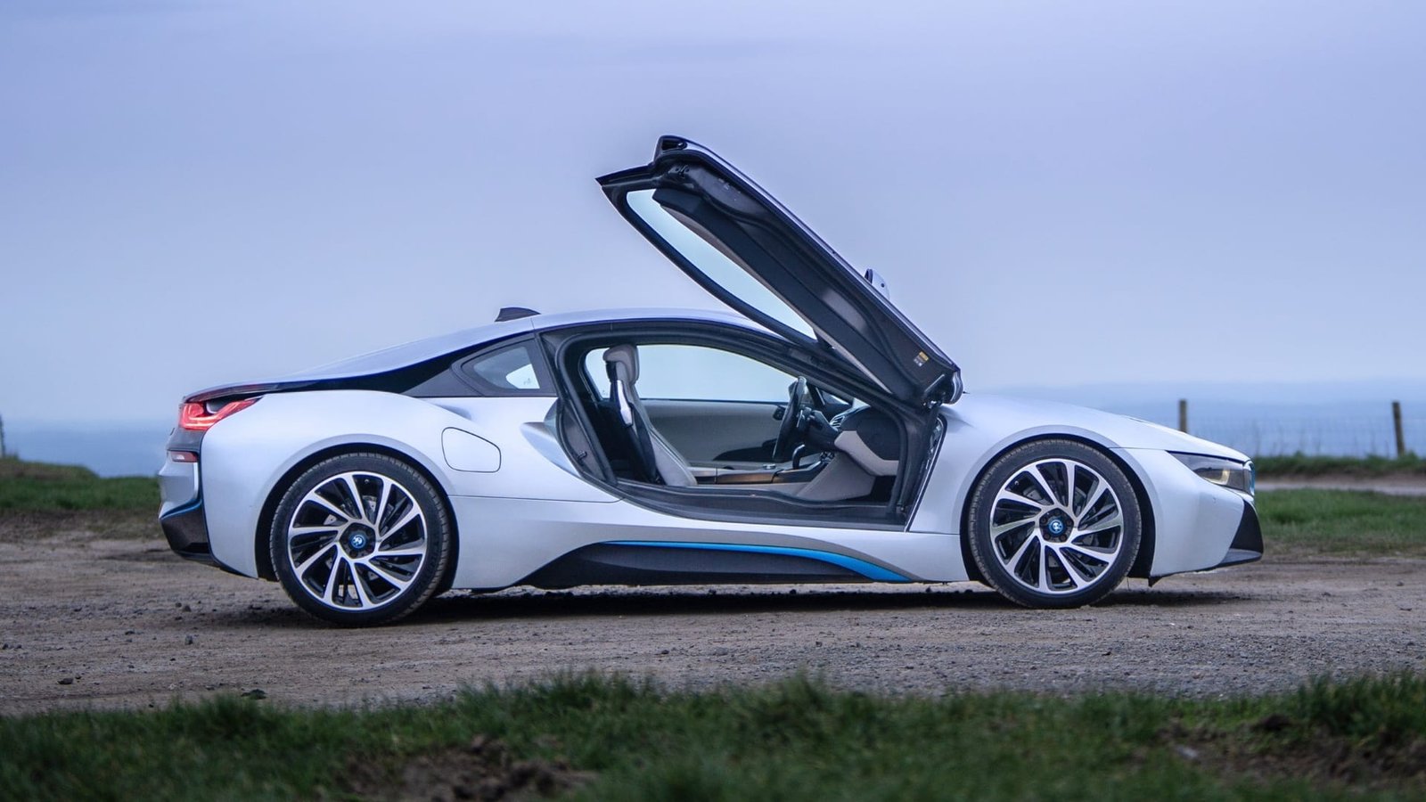 A silver BMW i8 with its butterfly door raised, parked on a gravel track above the sea